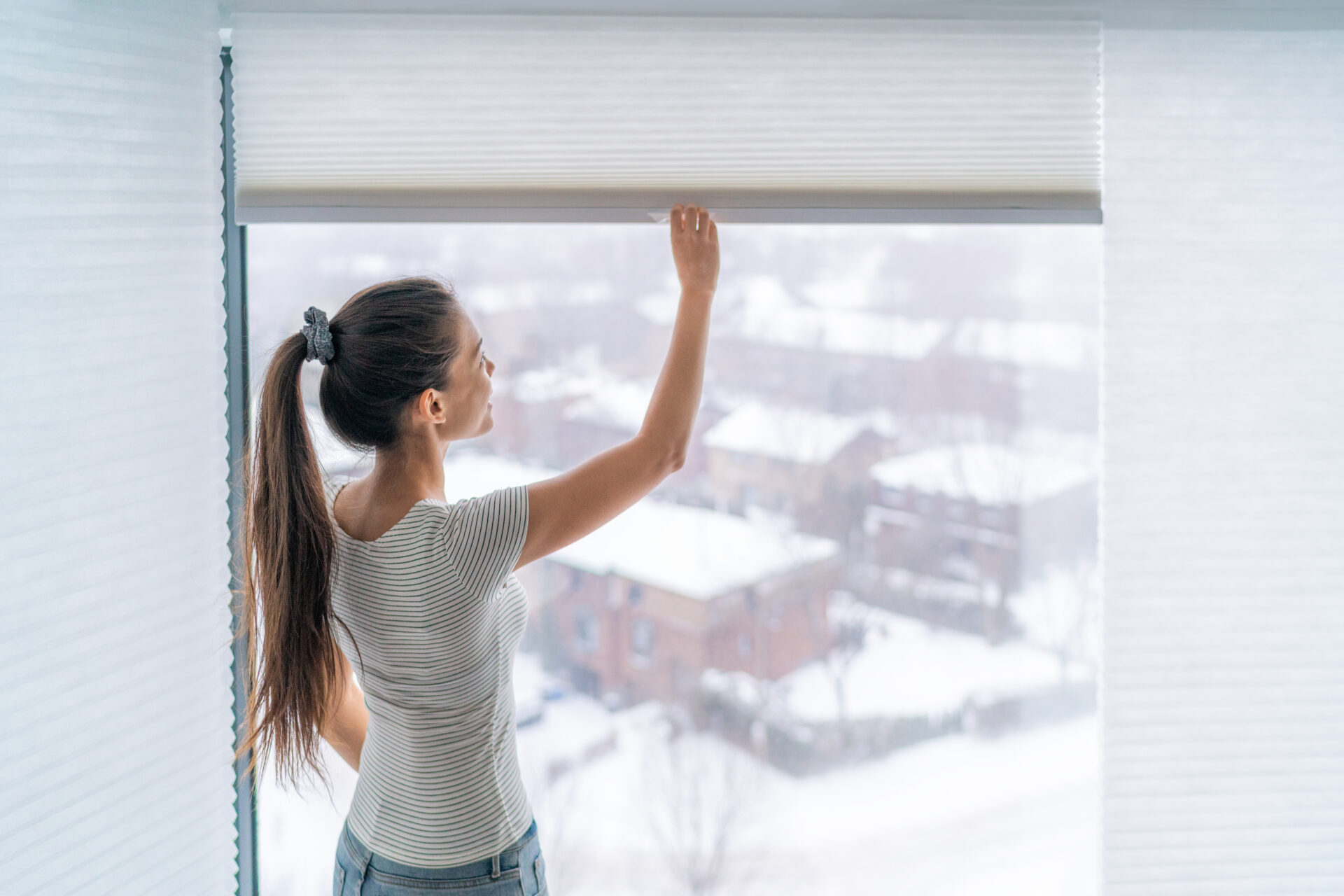 Woman opening cellular shades during winter morning. Find shades at Wholesale Blind Factory.